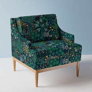 Op de afbeelding: Een groene fauteuil met een bloemenpatroon en houten poten. De stof heeft een verscheidenheid aan groene, blauwe, roze en gele bloemen en bladeren.