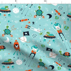 Op de afbeelding: Een turquoise blauwe stof met een herhalend patroon van ruimtethema-illustraties. Het patroon bevat katten, schildpadden, raketten, planeten, sterren en manen. De katten en schildpadden dragen astronautenhelmen. De raketten zijn rood en oranje met witte vlammen. De planeten zijn blauw en groen. De sterren zijn geel. De manen zijn wit. De stof is perfect voor een kinderkamer of een kinderkamer.