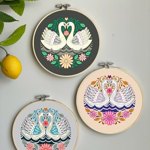 May include: Three embroidery hoops with a folk art design of two white swans facing each other with their necks intertwined. The swans are surrounded by flowers and leaves. The hoops are on a white background.