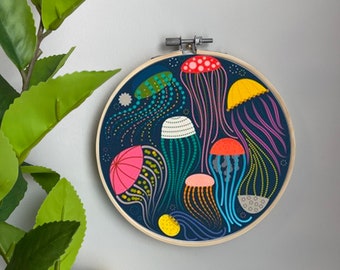 May include: A circular embroidery hoop with a dark blue background featuring colorful jellyfish with various patterns. The jellyfish are in different sizes and have long tentacles. The hoop is made of wood and has a natural finish.