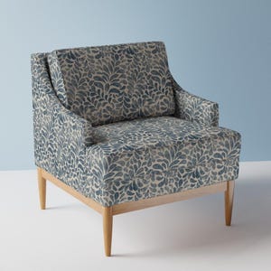 Pode incluir: Uma poltrona estofada em tecido azul e branco com um padrão repetitivo de folhas ou gotas. A cadeira possui um encosto quadrado, apoios de braços integrados e uma almofada de assento, tudo suportado por quatro pernas de madeira clara.