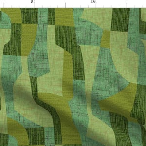 May include: Abstract fabric pattern in shades of green, olive, and teal. The design features geometric shapes and textured lines, creating a retro, mid-century modern aesthetic. The fabric appears to be a woven material, suitable for upholstery or home decor projects.