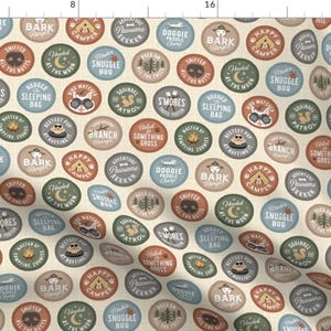 Op de afbeelding: Een beige stof met een herhalend patroon van kleurrijke ronde vlekken. Elke vlek heeft een andere camping-gerelateerde zin, zoals "Happy Camper", "Snuggle Bug" en "Bark Ranger".