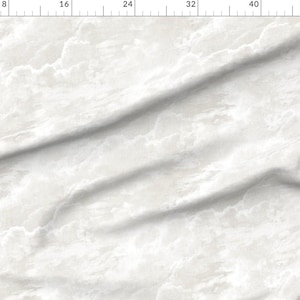 May include: A white fabric featuring a soft, cloudy pattern with subtle light gray and off-white tonal variations. The material is draped in diagonal folds, creating a textured appearance across the surface, with a ruler visible along the top edge.