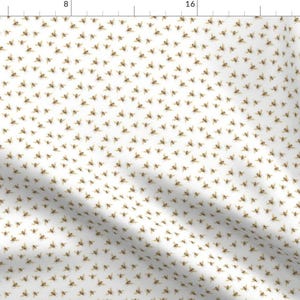 Op de afbeelding: Witte stof met een herhalend patroon van kleine, goudkleurige bijen. De bijen zijn in een verspringend patroon gerangschikt.