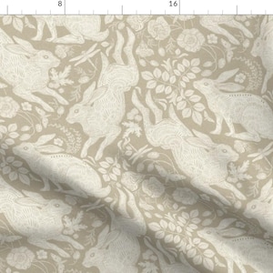Può includere: Tessuto beige con un motivo ripetuto di lepri bianche, fiori e fogliame. Le lepri sono raffigurate in varie pose, alcune che saltano e altre che riposano tra gli elementi floreali. Il design ha un aspetto vintage e botanico.