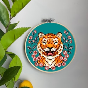 May include: Embroidered hoop art featuring a smiling tiger with orange and brown fur, surrounded by pink flowers and green leaves on a teal background.