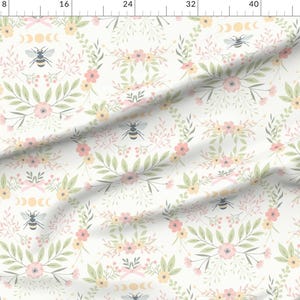 Pode incluir: Um tecido branco com um padrão repetido de flores rosa e amarelas, folhas verdes e abelhas com símbolos de lua e sol.