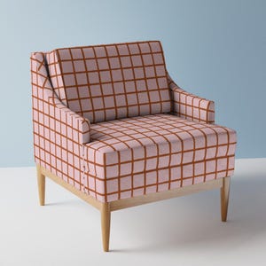 May include: An armchair upholstered in light pink fabric featuring a dark orange grid pattern. The chair has a square backrest, integrated armrests, and a seat cushion, all supported by four light-coloured wooden legs with a tapered design.