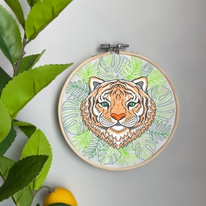 May include: Embroidered hoop art featuring a tiger face surrounded by green tropical leaves. The tiger has orange and black stripes, green eyes, and a pink nose.