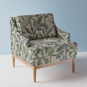 Könnte beinhalten: Gepolsterter Sessel mit hellem Holzrahmen. Der Stuhl hat ein botanisches Muster mit grünen Blättern, blauen Akzenten und braunen Ästen auf neutralem Hintergrund. Der Stuhl hat eine hohe Rückenlehne und Armlehnen.