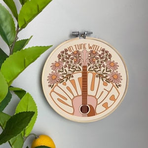 May include: A round embroidery hoop with a beige background featuring a pink acoustic guitar with floral accents. The text "In tune with nature" is written in a retro font around the guitar.