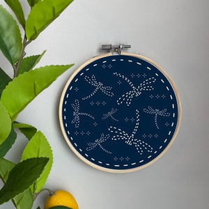 Könnte beinhalten: Ein marineblauer Stickrahmen mit weißen Stichen, der ein Muster aus Libellen darstellt. Der Rahmen ist aus Holz und hat eine natürliche Oberfläche.