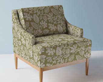 May include: An upholstered armchair with a light wood frame. The chair features a green and cream-colored leaf pattern fabric. The chair has a high back, armrests, and four tapered legs. The background is a light blue.