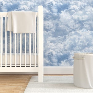 May include: A white cot with a white blanket on it, a white laundry basket, and a white rug in front of a wall with a blue and white cloud wallpaper.