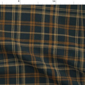 May include: A woven fabric featuring a plaid pattern with dark navy blue and warm tan brown intersecting lines. The textile is displayed against a ruler showing measurements in inches, with the pattern repeating across the surface.
