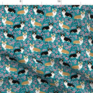 Op de afbeelding: Een tealblauwe stof met een herhalend patroon van corgi-honden en roze kersenbloesems. De corgis zijn in drie kleurvariaties: zwart-wit, bruin-wit en rood-wit.
