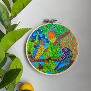 May include: A colorful embroidery hoop with a blue, green, orange, and purple bird design. The birds are perched on branches with green leaves and brown acorns.