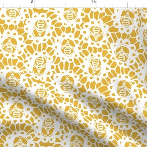 May include: A yellow fabric with a white lace pattern featuring bees. The bees are in a circle with wings spread out. The fabric is a repeating pattern.