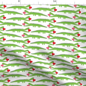 May include: A repeating pattern of green alligators holding red and pink hearts on a white background. The alligators are smiling, creating a playful and whimsical design. This fabric is suitable for various craft projects.
