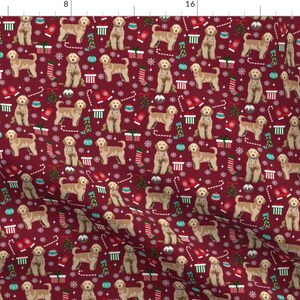May include: A red fabric with a repeating pattern of golden doodle dogs wearing Santa hats and surrounded by Christmas decorations like candy canes, snowflakes, and stockings.