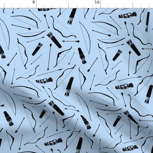 May include: Pale blue fabric featuring a repeating pattern of black bows, arrows, and quivers. The design includes various bow shapes and arrow styles, creating a dynamic and illustrative print. The fabric is suitable for crafting or apparel.