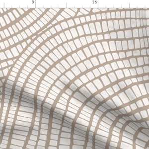May include: A patterned fabric featuring off-white rectangular shapes on a tan backdrop. The rectangles are arranged in curved, grid-like rows, creating a textured, organic design. The fabric is folded, showing depth and dimension.