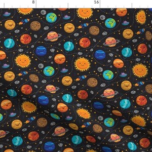 Puede incluir: Una tela negra con un patrón repetido de planetas, estrellas y cohetes. Los planetas son todos sonrientes y tienen diferentes colores. Las estrellas son amarillas y los cohetes son rojos.