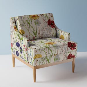 May include: An armchair upholstered in an off-white fabric featuring a colorful botanical print of red poppies, yellow dandelions, blue cornflowers, and various green grasses. The chair has a square back, low arms, and four light-toned wooden legs.
