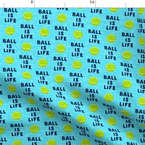 Op de afbeelding: Een blauwe stof met een herhalend patroon van gele tennisballen en de tekst "Ball is Life" in donkerblauw.