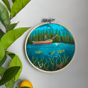 May include: A circular embroidery hoop with a blue background depicting a person fishing from a red boat in a lake. The scene is set at night with a full moon and stars in the sky. The water is clear and you can see fish swimming below the boat. The hoop is made of wood and has a natural finish.
