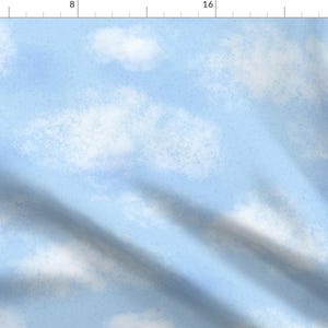 May include: A light blue fabric featuring a repeating pattern of soft, white, fluffy clouds. The textile displays a subtle, painterly texture throughout the sky-blue background, with a visible ruler along the top and left edges for scale.