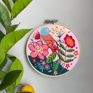 May include: An embroidery hoop featuring a pink and green floral design with a blue and orange bird perched on a branch. The hoop is surrounded by a wooden frame.