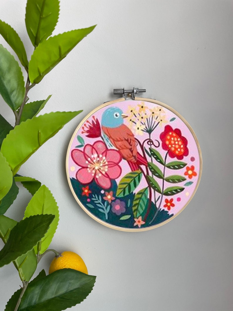 May include: An embroidery hoop featuring a pink and green floral design with a blue and orange bird perched on a branch. The hoop is surrounded by a wooden frame.