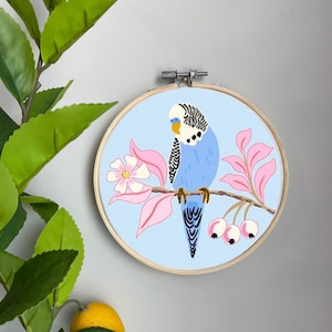 Könnte beinhalten: Ein blauer und weißer Sittich, der auf einem Zweig mit rosa Blüten und roten Beeren sitzt. Der Vogel ist auf einem hellblauen Hintergrund in einem runden Holzreifen gestickt.