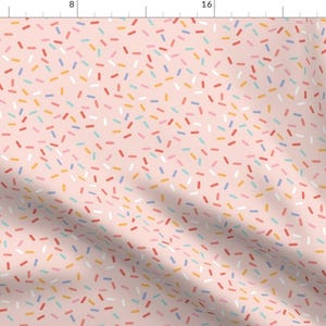 May include: A pink fabric with a repeating pattern of colorful sprinkles. The sprinkles are in various shades of red, orange, yellow, blue, and white.