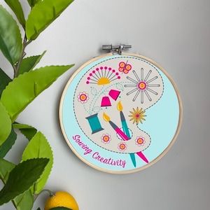 May include: A light blue embroidery hoop with a beige palette design featuring sewing and crafting tools. The text "Sewing Creativity" is written in pink on the hoop.