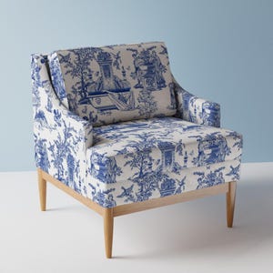 Puede incluir: Un sillón blanco con un patrón de toile azul y blanco que representa una escena de jardín. El sillón tiene un marco de madera con patas cónicas.