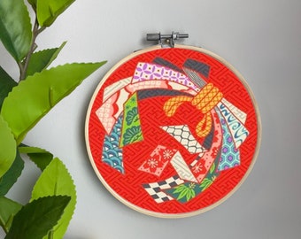 May include: A red embroidery hoop with a colorful Japanese-inspired design featuring geometric patterns and a yellow bow. The design is made up of various fabrics with different textures and patterns.