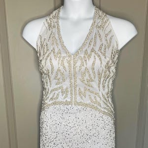 May include: White, floor-length formal dress with a V-neck halter top. The bodice features a beaded design, and the skirt is covered in sequins. The dress is displayed on a white mannequin.