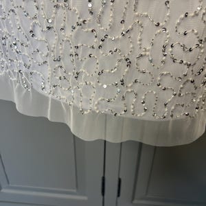 May include: Close-up of a white garment with detailed embellishments. The fabric is decorated with a delicate pattern of silver sequins and small white beads, creating a shimmering effect. The hemline has a soft, ruffled edge.