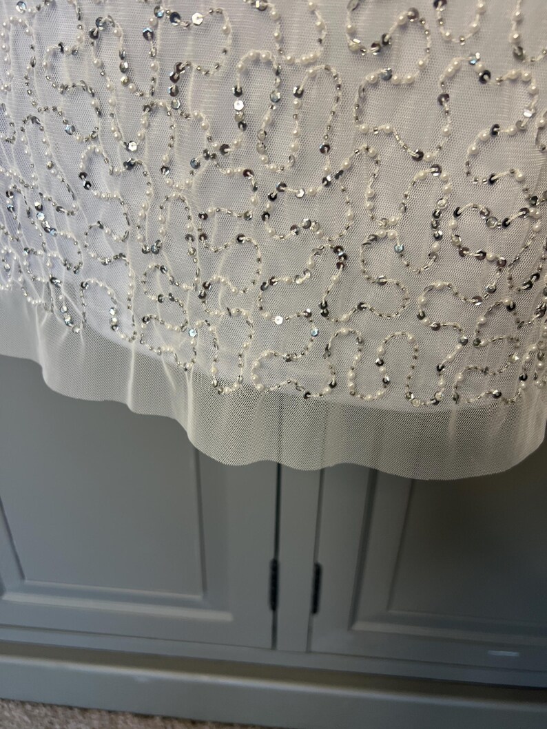 May include: Close-up of a white garment with detailed embellishments. The fabric is decorated with a delicate pattern of silver sequins and small white beads, creating a shimmering effect. The hemline has a soft, ruffled edge.