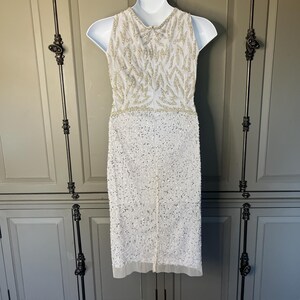 May include: A white sleeveless dress adorned with detailed beadwork. The bodice has a pattern of gold beads, and the skirt is covered in white sequins. The dress is displayed on a white mannequin, highlighting its elegant design and craftsmanship.