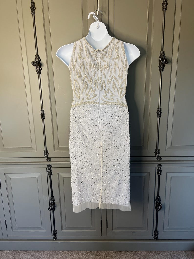 May include: A white sleeveless dress adorned with detailed beadwork. The bodice has a pattern of gold beads, and the skirt is covered in white sequins. The dress is displayed on a white mannequin, highlighting its elegant design and craftsmanship.