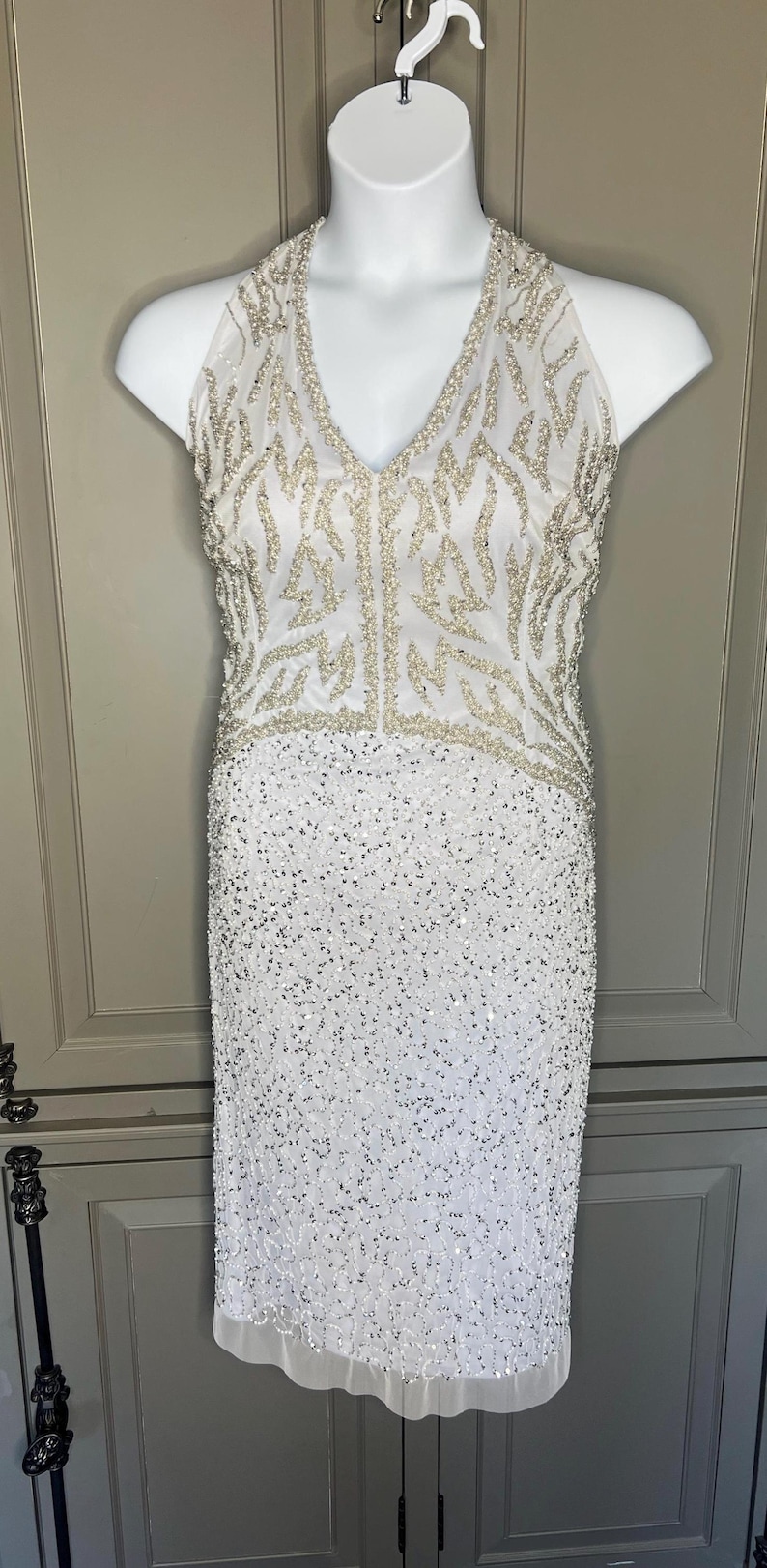 May include: White, floor-length formal dress with a V-neck halter top. The bodice features a beaded design, and the skirt is covered in sequins. The dress is displayed on a white mannequin.