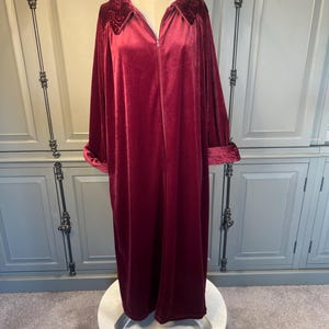 May include: A full-length, burgundy velvet robe with a zip-up front and a wide collar. The collar and cuffs feature a darker, textured velvet detail. The robe is displayed on a mannequin, showcasing its flowing design and luxurious fabric.