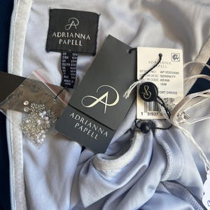 May include: A light blue Adrianna Papell dress with tags attached. The tags display the brand logo, size, and style information. A small bag of beads is also visible. The dress is a size 22W.