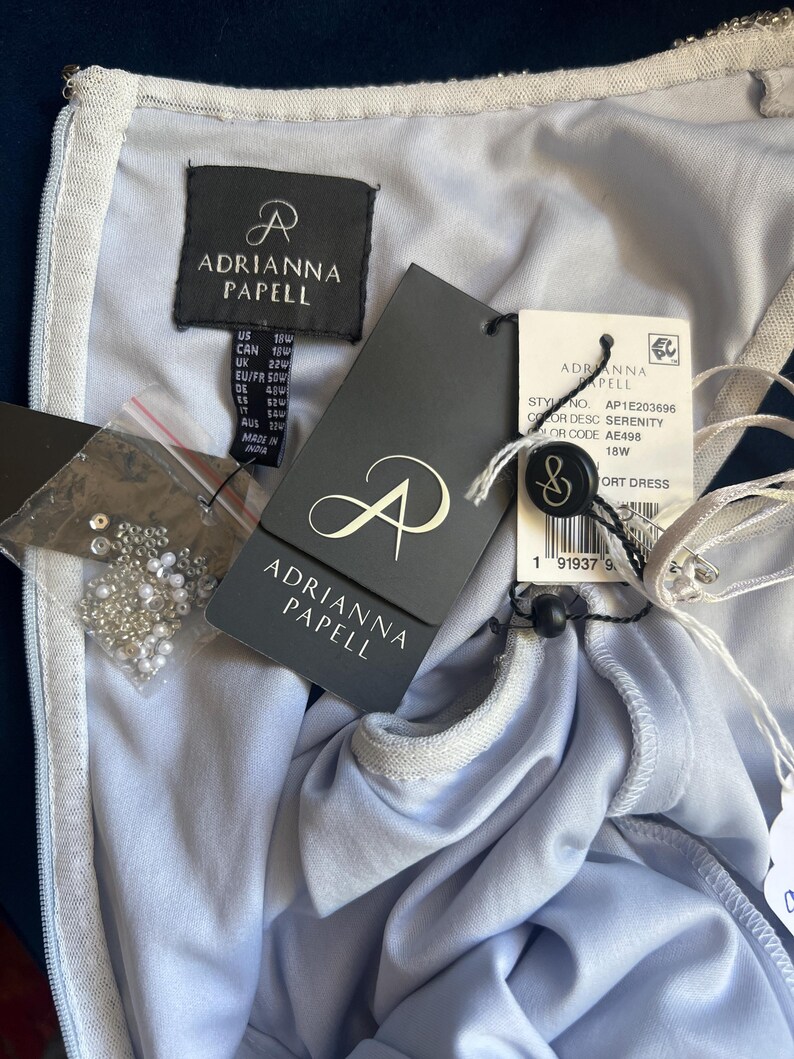 May include: A light blue Adrianna Papell dress with tags attached. The tags display the brand logo, size, and style information. A small bag of beads is also visible. The dress is a size 22W.