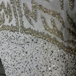 May include: Close-up of a white garment with detailed embellishments. The design incorporates silver beading and sequins in flowing patterns. Small, round pearls are interspersed, adding texture and dimension to the fabric.