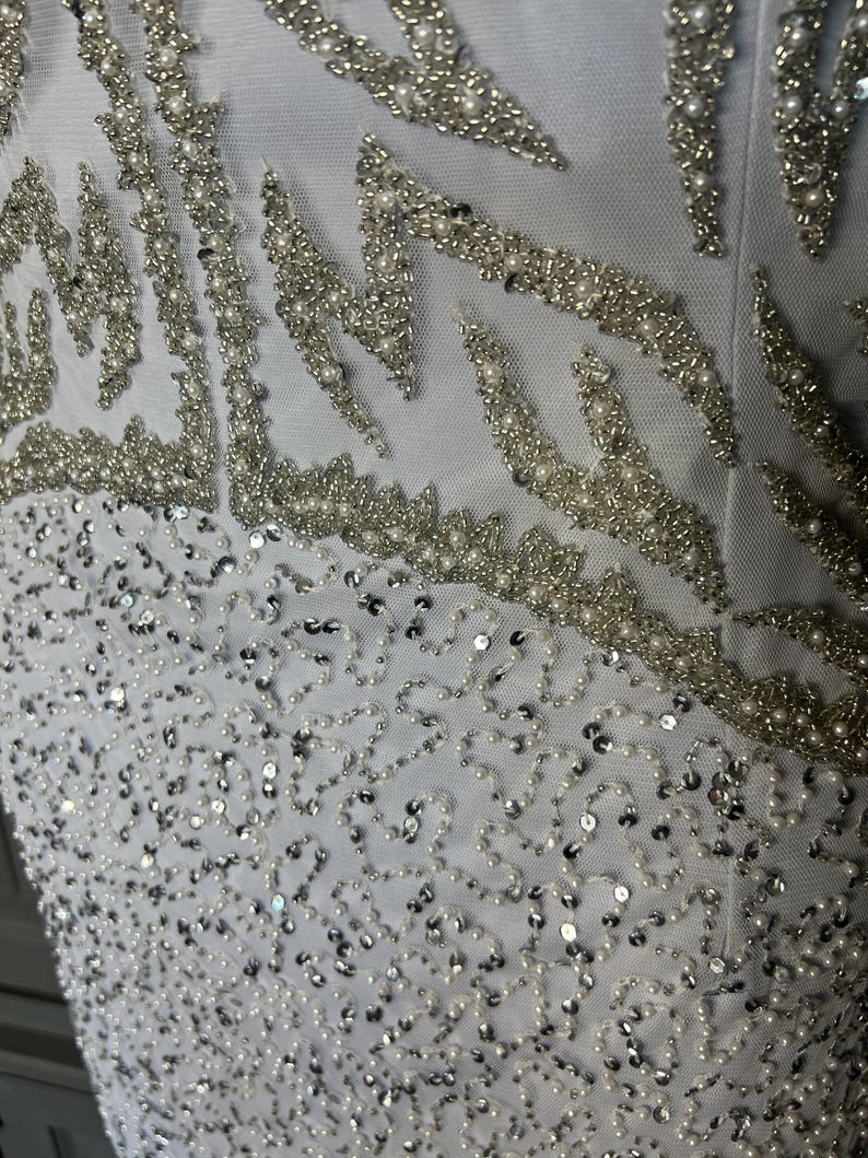 May include: Close-up of a white garment with detailed embellishments. The design incorporates silver beading and sequins in flowing patterns. Small, round pearls are interspersed, adding texture and dimension to the fabric.